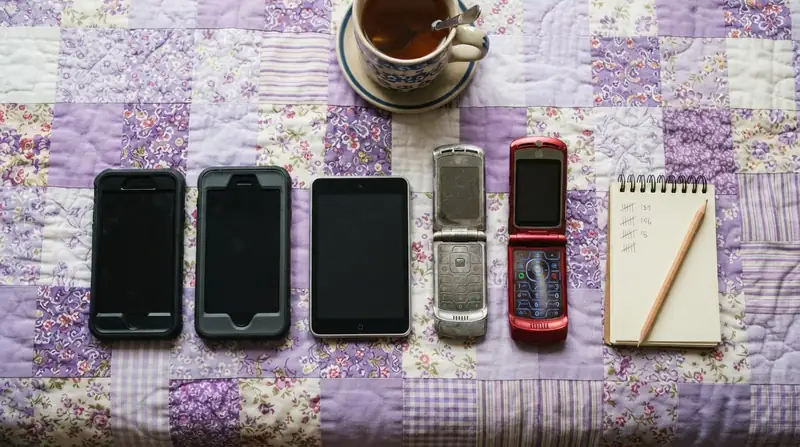 Five different phones laid in a row on a lavender quilt with a notepad and pencil, a red flip phone at the end